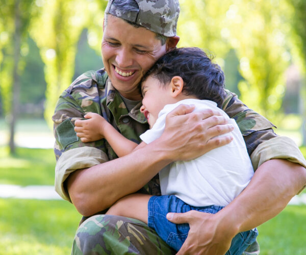 Cheerful military dad hugging little son, holding boy In arms outdoors after returning from mission trip. Vertical shot. Family reunion or returning home concept